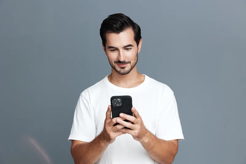 Full body smiling young man wearing white t-shirt looking at smartphone, standing against grey background. Technology and lifestyle concept.