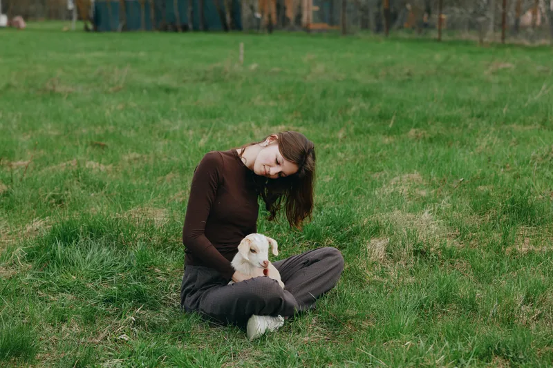 Cheerful woman outdoors, sitting on grass with a playful goat kid, gentle touch, natural light, serene meadow atmosphere, candid moment, rustic vibe, soft colors in green field under clear sky,