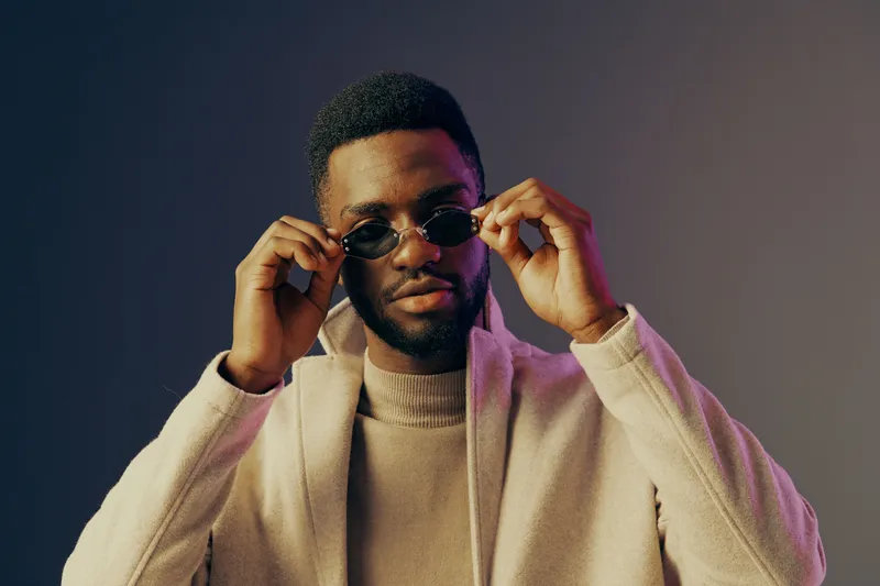 Stylish African man in sunglasses and a light sweater, posing confidently against a gradient background, showcasing modern urban fashion and self expression