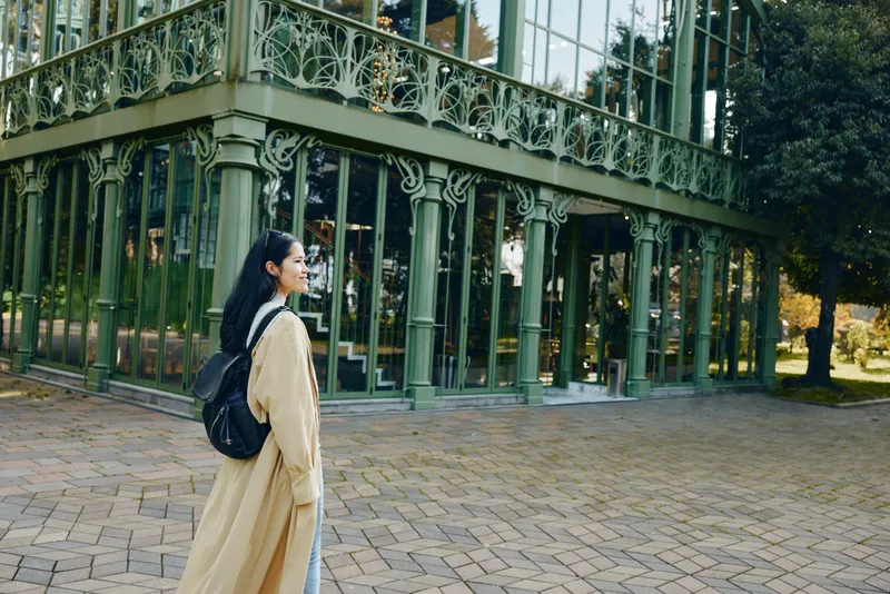 Woman with backpack stands beside a glass conservatory, long beige coat in autumn light, and tranquil courtyard scene, blending architecture, outdoor aesthetics and calm travel mood.