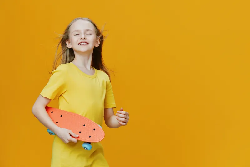 Happy girl with long hair and yellow dress holding a skateboard against a bright orange background, conveying joy, fun, and youthfulness in a vibrant setting