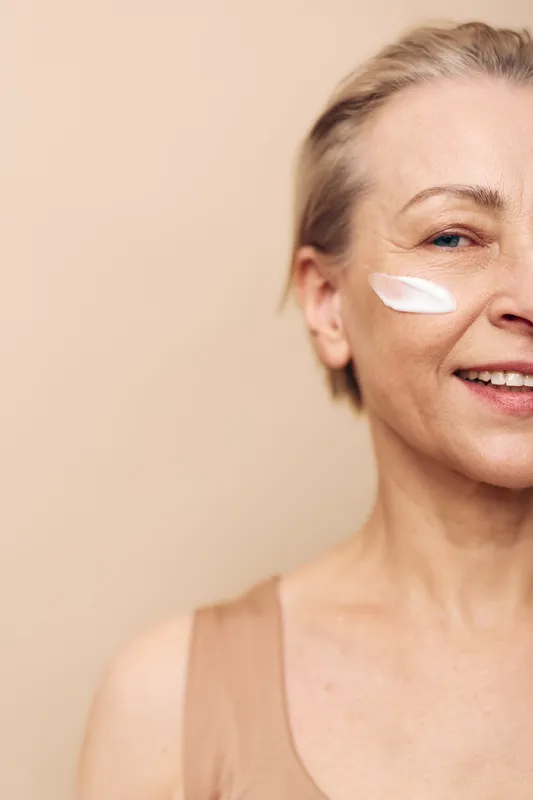 Smiling mature woman applies white cream on cheek, demonstrating skincare routine for aging skin. Close-up on beige background with natural light. Concept of beauty, wellness, and self-care.