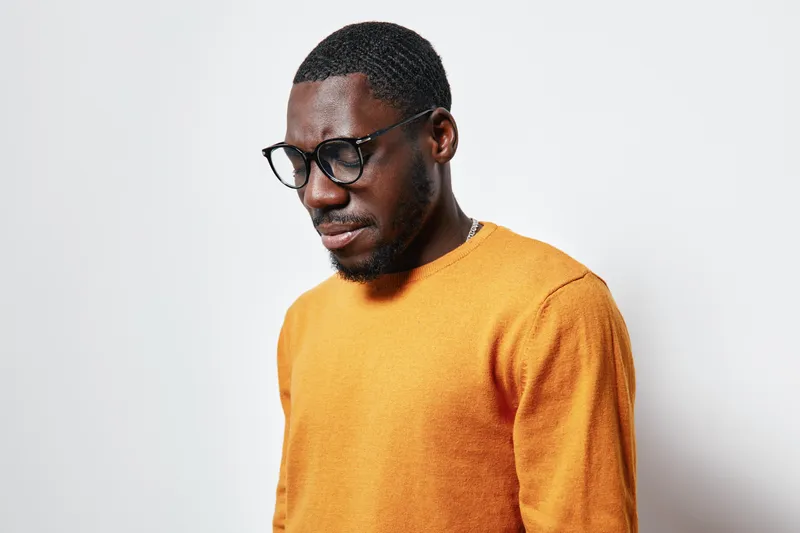 African American man with dark skin wearing glasses and orange sweater standing against white background looking down thoughtfully. Casual portrait with natural light and minimalistic style.