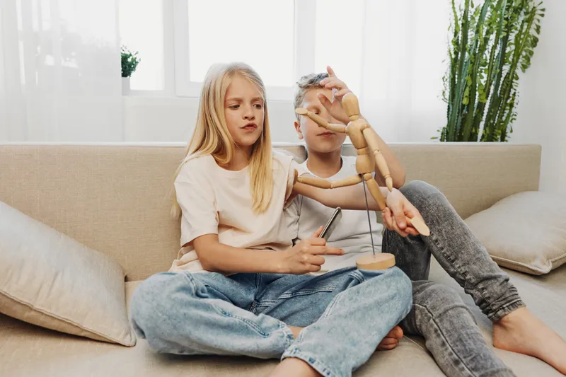 Two children sitting on sofa playing with wooden mannequin, focused on creativity and learning in cozy bright living room environment.