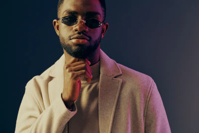 Stylish young African American man in sunglasses wears a beige coat against a dark gradient background, exuding confidence and modern fashion