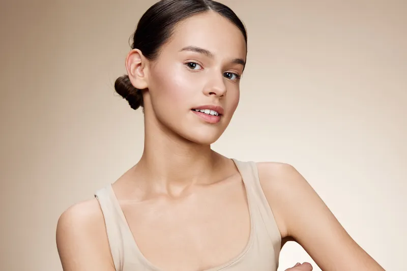 Natural beauty portrait of a young woman with a soft expression, wearing minimal makeup, showcasing flawless skin against a neutral background