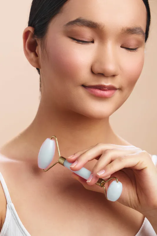 asian woman gently uses a facial roller in a soft, clean setting, smiling with calm confidence as she practices self care and skincare routine today, mindful and serene.
