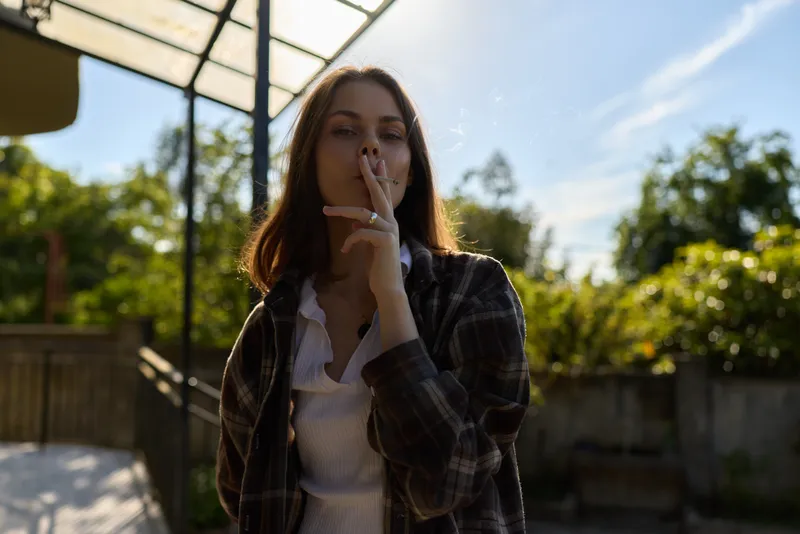 A young woman in a plaid shirt stands outdoors, smoking softly and smiling faintly, delivering a candid lifestyle moment with a relaxed street vibe