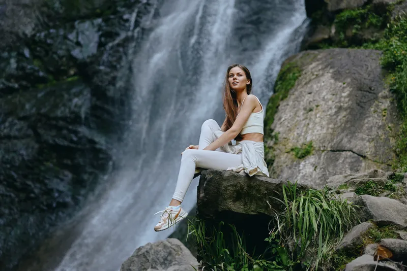 young woman sitting on rock near waterfall in outdoor nature setting, wearing white casual clothes, relaxed and thoughtful mood, vibrant greenery background