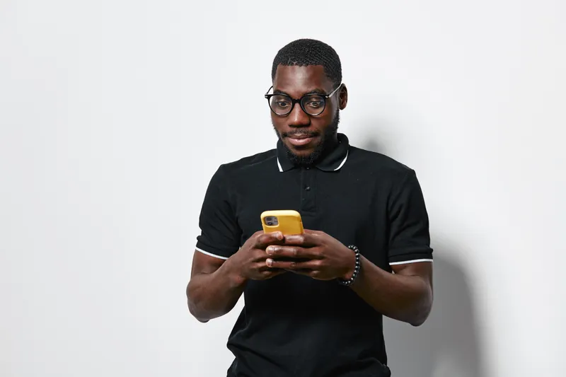 Black man with dark skin wearing glasses and black polo shirt using yellow smartphone standing against white background. Casual technology and communication concept.