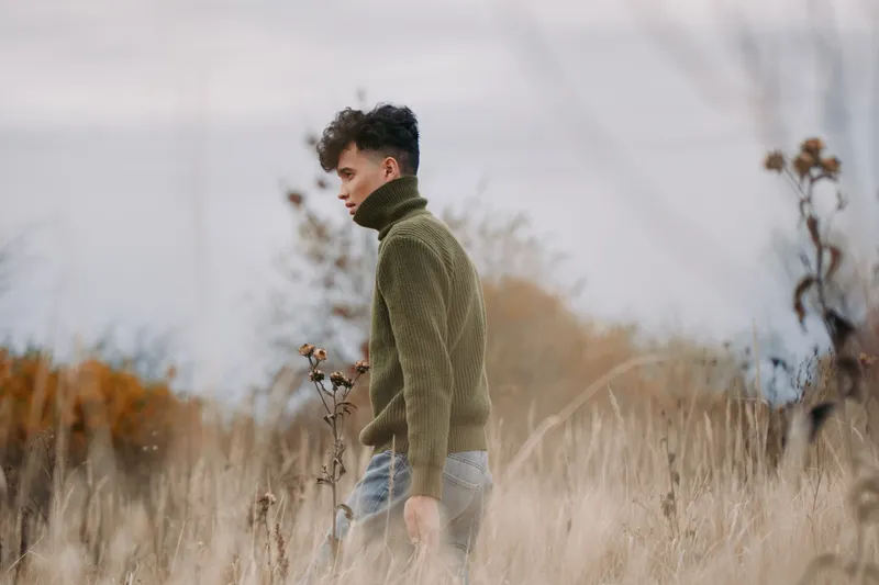 Young man walks through tall autumn grass wearing a knit turtleneck, captured in an authentic, understated moment conveying natural presence and credible candid portrait mood.