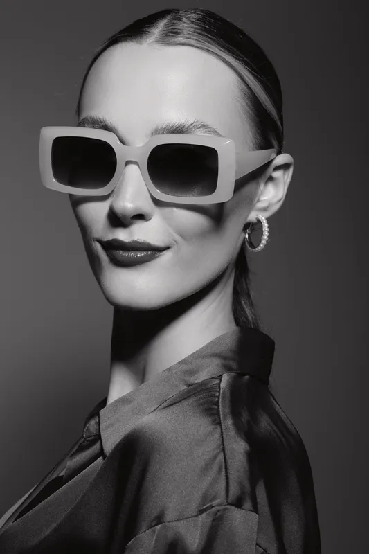 Stylish white woman with pale skin wearing large square sunglasses and hoop earrings smiles softly in black and white close-up studio portrait. Fashion concept.