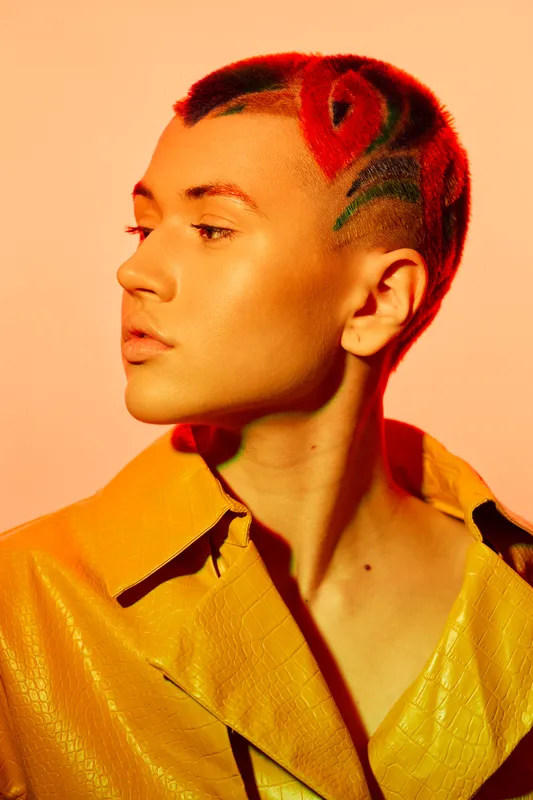 Young woman with artistic short hairstyle and vibrant red flower design, wearing a bold yellow textured jacket against a warm orange background.