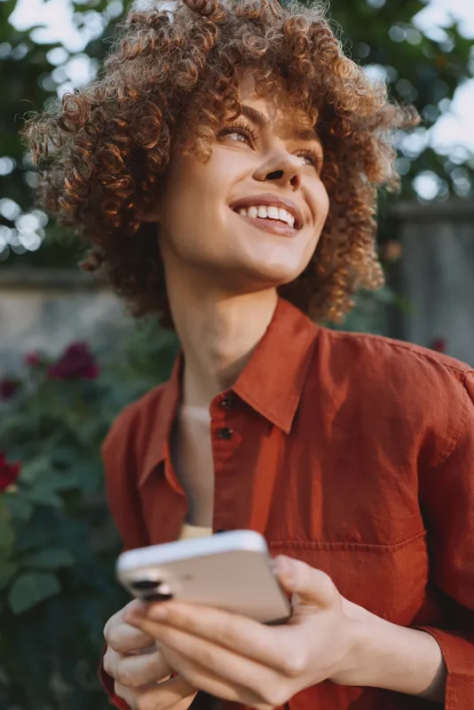 Woman lifestyle outdoor portrait, smiling and relaxed, holding smartphone, casual red shirt, natural light, garden backdrop, candid moment of everyday joy and connection