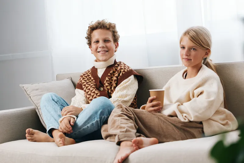 Two children sit on a light sofa wearing cozy sweaters, relaxed and smiling, as natural daylight fills a warm living room, suggesting comfort, family, and candid everyday life.