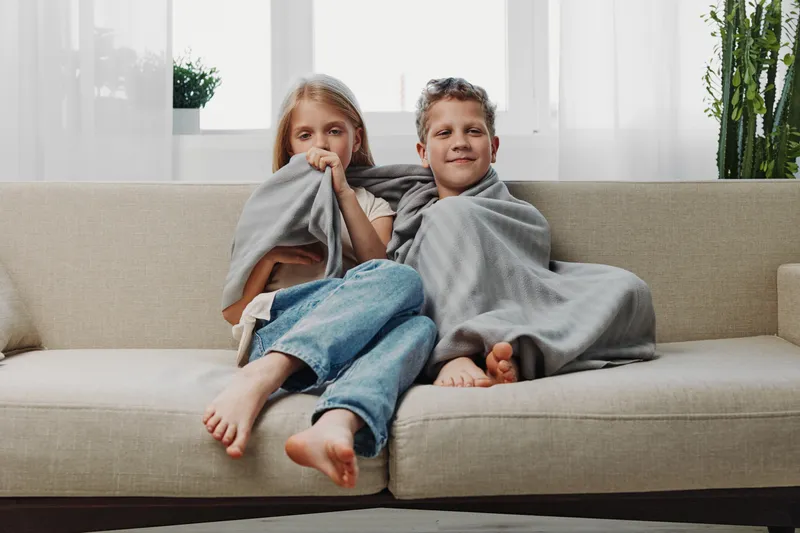 Two children wrapped in gray blankets sit barefoot on beige sofa in bright room, cozy and relaxed atmosphere at home with natural light.