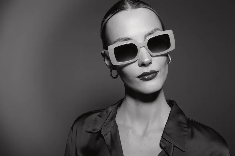Stylish white woman with pale skin in oversized sunglasses poses confidently in black shirt. Monochrome portrait with high contrast lighting and fashion concept.