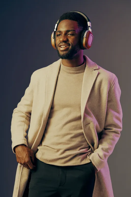 Stylish young Black man wearing headphones and a beige coat, smiling against a gradient background, expressing joy and confidence