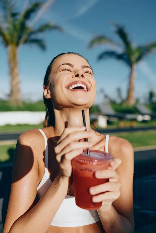 Happy young woman in sportswear enjoys a cold red smoothie outdoors under palm trees, smiling with eyes closed, showing healthy lifestyle and summer relaxation mood.