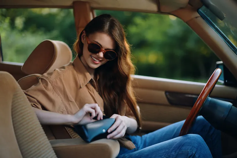 Smiling young woman in sunglasses enjoying summer drive, wearing casual outfit, with nature background and stylish interior, emphasizing freedom and leisure.