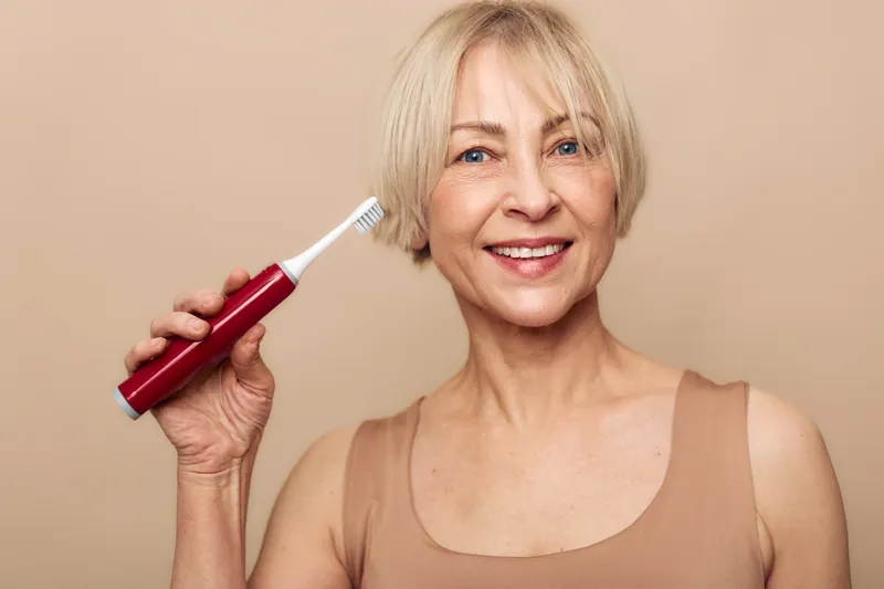 Smiling mature woman with short blonde hair holds red electric toothbrush near her face against beige background. Concept of dental hygiene, oral care, healthy lifestyle, and personal wellness.