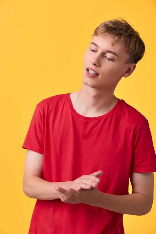 Young man in a red shirt demonstrates a gesture with hands and an expressive face against a yellow background, conveying casual energy, emotion, and lifestyle content