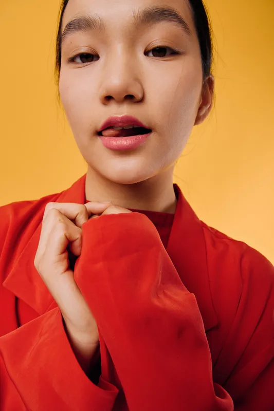 Young woman wearing bright red jacket posing with hand close to collar against yellow background in studio. Fashion portrait with serious expression and minimal makeup on face.