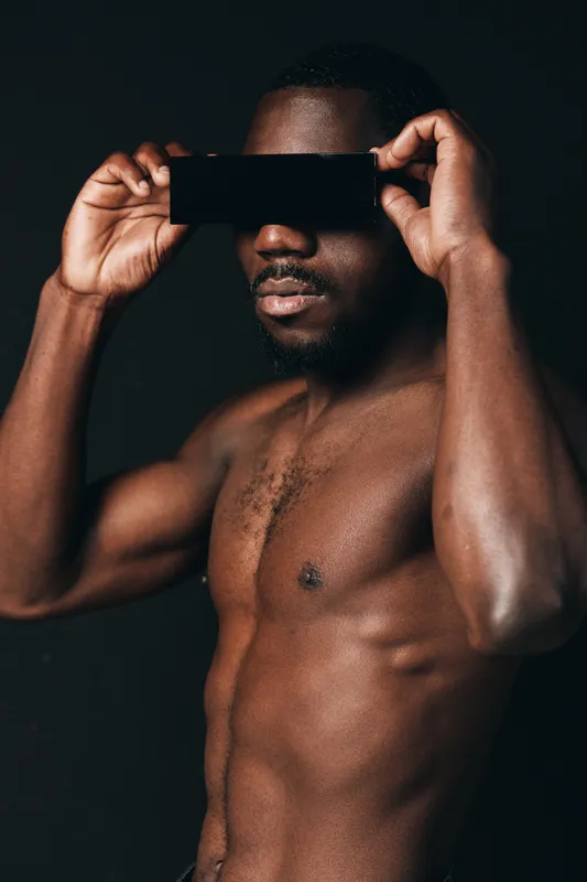 Black man with dark skin holding black card in front of eyes in creative conceptual visual style. Portrait of shirtless African male with muscular body on dark background for artistic expression.