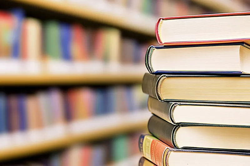Аккуратно сложенная стопка книг разных цветов на блестящей поверхности на фоне библиотеки с мягким фокусом, заполненной бесчисленными книгами