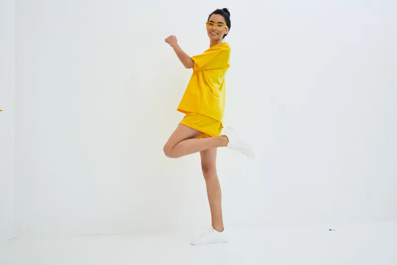 young woman in yellow outfit jumping joyfully in bright studio with white background, wearing glasses and white sneakers, expressing happiness and energy