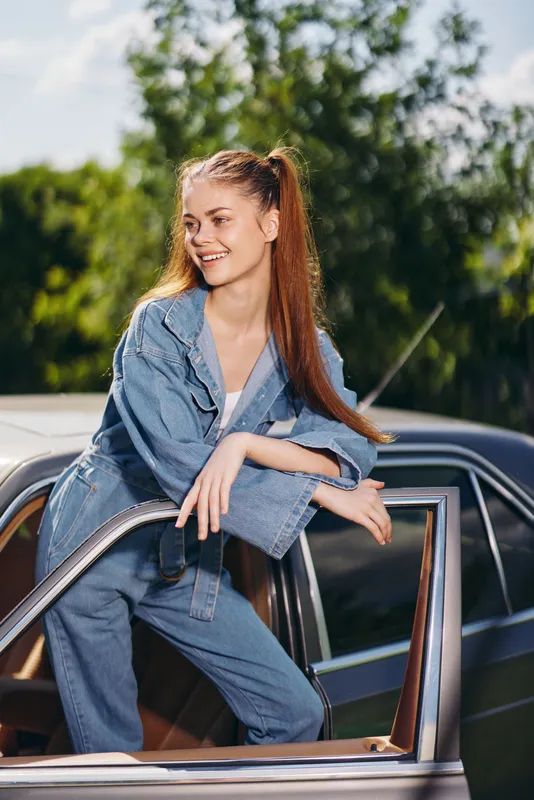 Young woman in denim outfit smiling by classic car against green background, capturing summer vibes and carefree emotions, perfect for lifestyle photography.