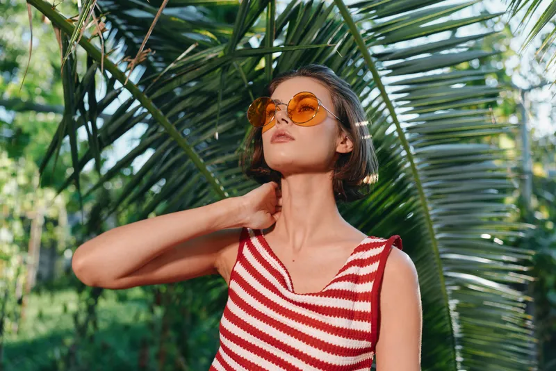 A cheerful woman wearing a red and white striped sleeveless top stands outdoors among tropical plants. She wears round sunglasses and looks into the distance with a relaxed smile, creating a sunny,