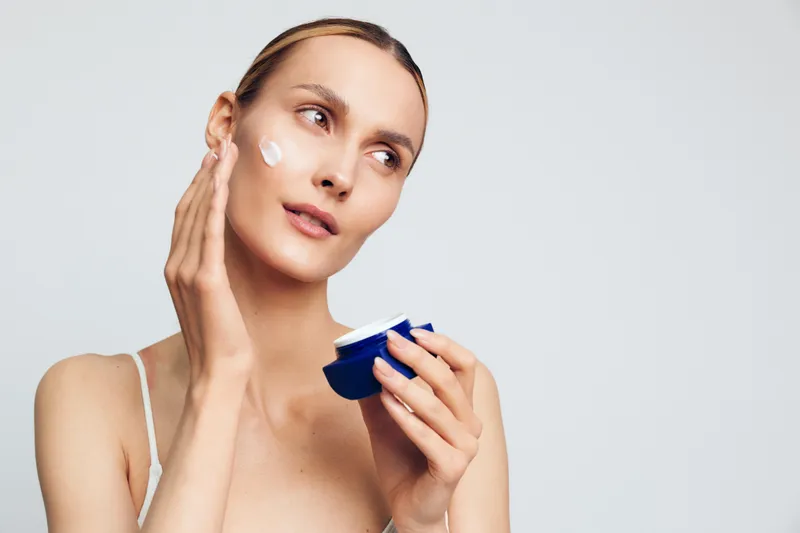A young woman with clear skin applies white cream on her cheek using fingers while holding a blue jar in a bright studio. Concept for skincare, beauty, and self-care routine.