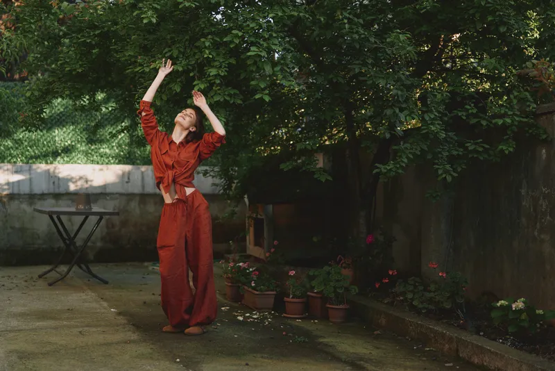 A young woman in a red outfit dances joyfully in a sunlit garden courtyard, arms raised above her head, surrounded by potted plants and lush green foliage