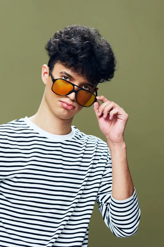 Young man wearing striped shirt and orange sunglasses, posing confidently against olive background with curly hair and casual style.