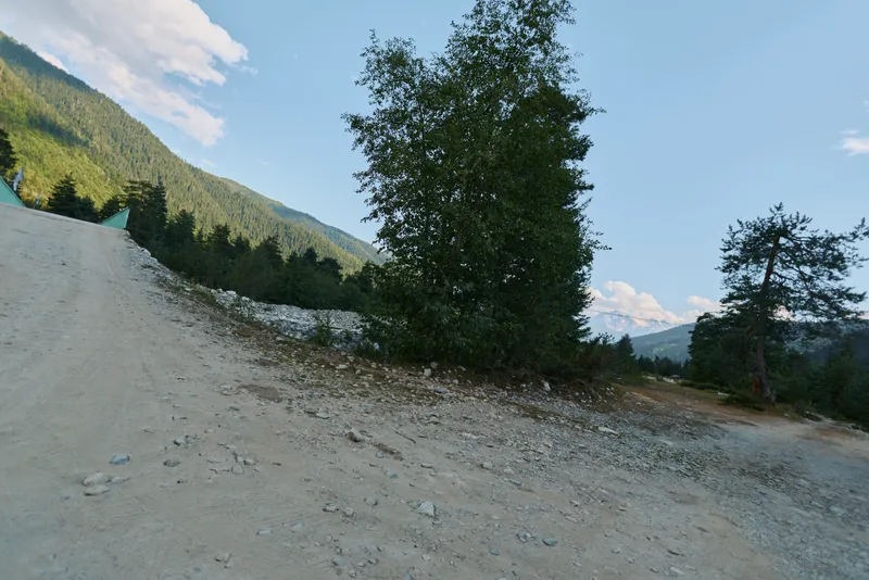 A rugged mountain road winds through a rocky landscape under a bright blue sky. Sparse trees line the track, creating a sense of solitude and outdoor exploration.