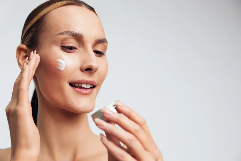 Young woman applying face cream with fingers, skincare routine, natural beauty, clear skin, soft lighting, closeup portrait, white background, healthy lifestyle concept