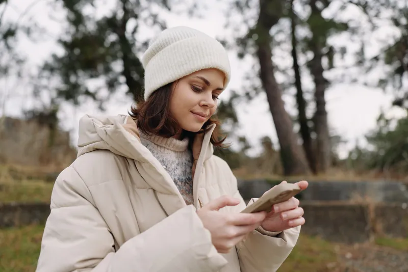 Woman in a warm coat and beanie using a smartphone outdoors in a park, focused expression, chilly weather, casual tech use and nature backdrop