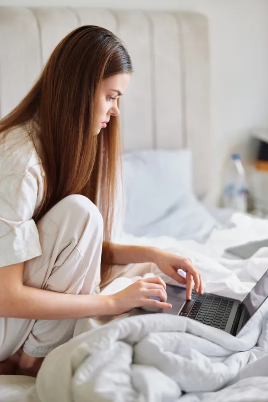 Woman on bed using laptop in bedroom, working remote in pajamas at home. Cozy morning scene with focused freelancer typing on laptop, casual work from home routine and calm focus.