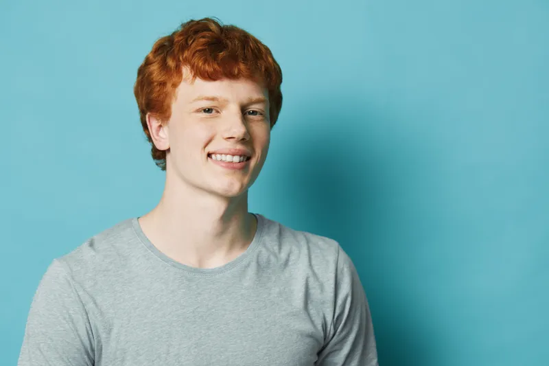 Young man with red hair smiling in casual gray shirt against blue background. Portrait of cheerful male with freckles and short hairstyle, relaxed and confident expression in studio.