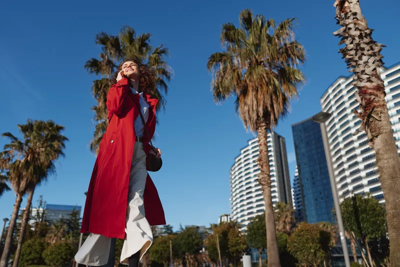 A woman in a red trench coat walks outdoors among palm trees and modern buildings, looking up with a confident smile and breezy motion.