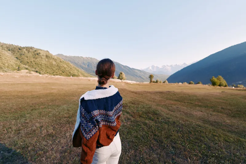 Woman back in cozy sweater stands in open meadow facing distant mountains and wide landscape, solitary traveler enjoying nature, autumn light and peaceful outdoor scenery