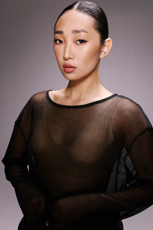 Модная азиатская женщина в прозрачном чёрном топе позирует на сером фоне для фотосессии