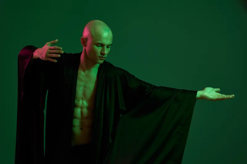 A man in a flowing black robe poses dramatically against a green backdrop, showcasing strength and charisma. The lighting creates a striking, artistic atmosphere.