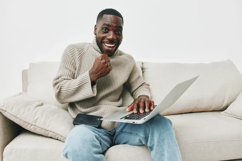 Cheerful African man in cozy sweater expressing excitement while using a laptop at home, surrounded by a minimalist light colored interior