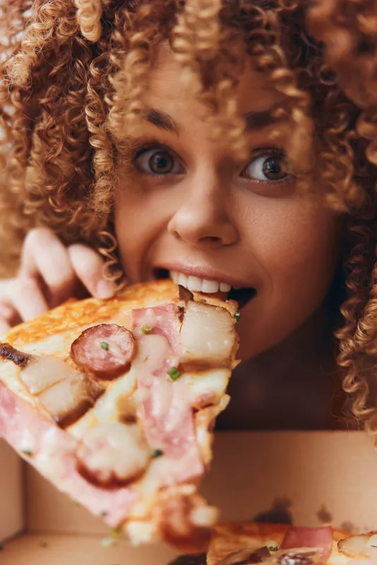 Кудрявая женщина наслаждается кусочком пиццы из коробки, в которой не хватает одного куска
