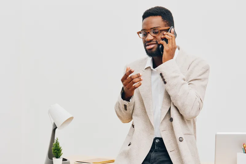 Confident smiling man with glasses in beige blazer talking on mobile phone while standing at workplace with modern desk and minimalistic decor. Business communication, professional lifestyle concept.
