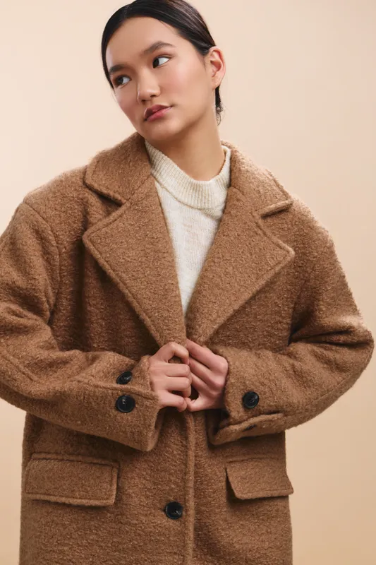Asian woman wears a cozy brown coat in a soft studio setting, gazing thoughtfully as she adjusts her lapels, creating a calm stylish mood.