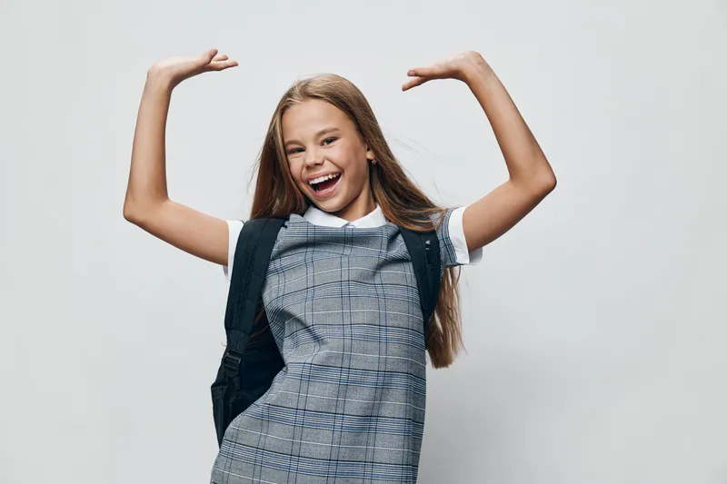 Happy girl smiling and posing with raised arms in checkered dress and backpack, isolated on gray background with joyful expression and long hair for childhood portrait.