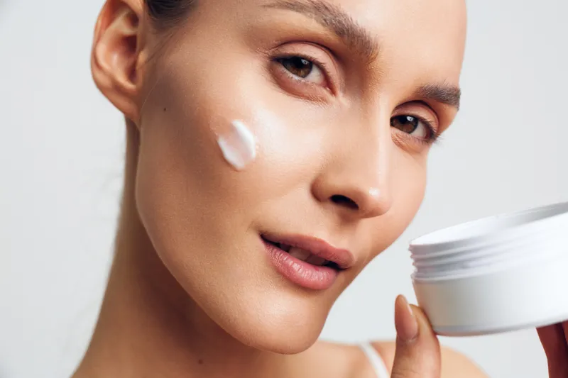 Young woman applies moisturizing cream on her face closeup holding white jar skincare routine natural beauty healthy skin care concept isolated on white background.